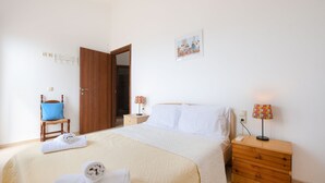 A neatly made bed with white linens, a wooden nightstand with a lamp, a chair with a blue cushion, and a door to another room. - Angelika House -Amazing Sea view (Faistos)