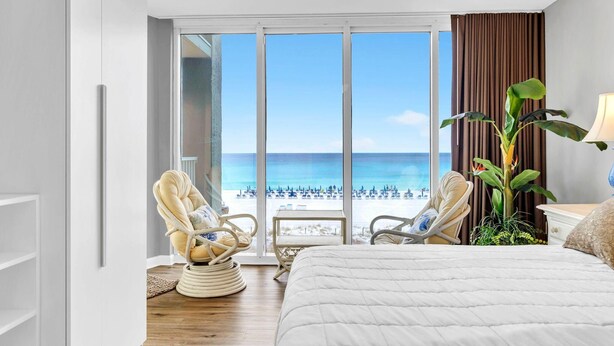 Een hotelkamer met een groot bed, twee stoelen, een plant en uitzicht op het strand.
