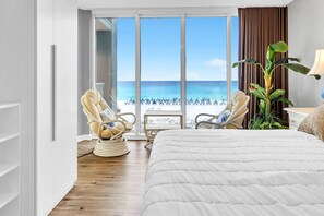 Une chambre d’hôtel avec un grand lit, deux fauteuils, une plante et une vue sur la plage.
