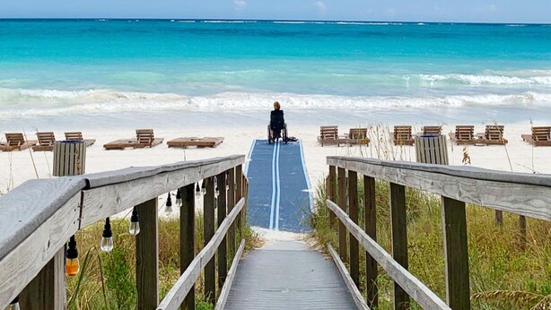A wooden boardwalk leading to a beach with lounge chairs and a person in a wheelchair.