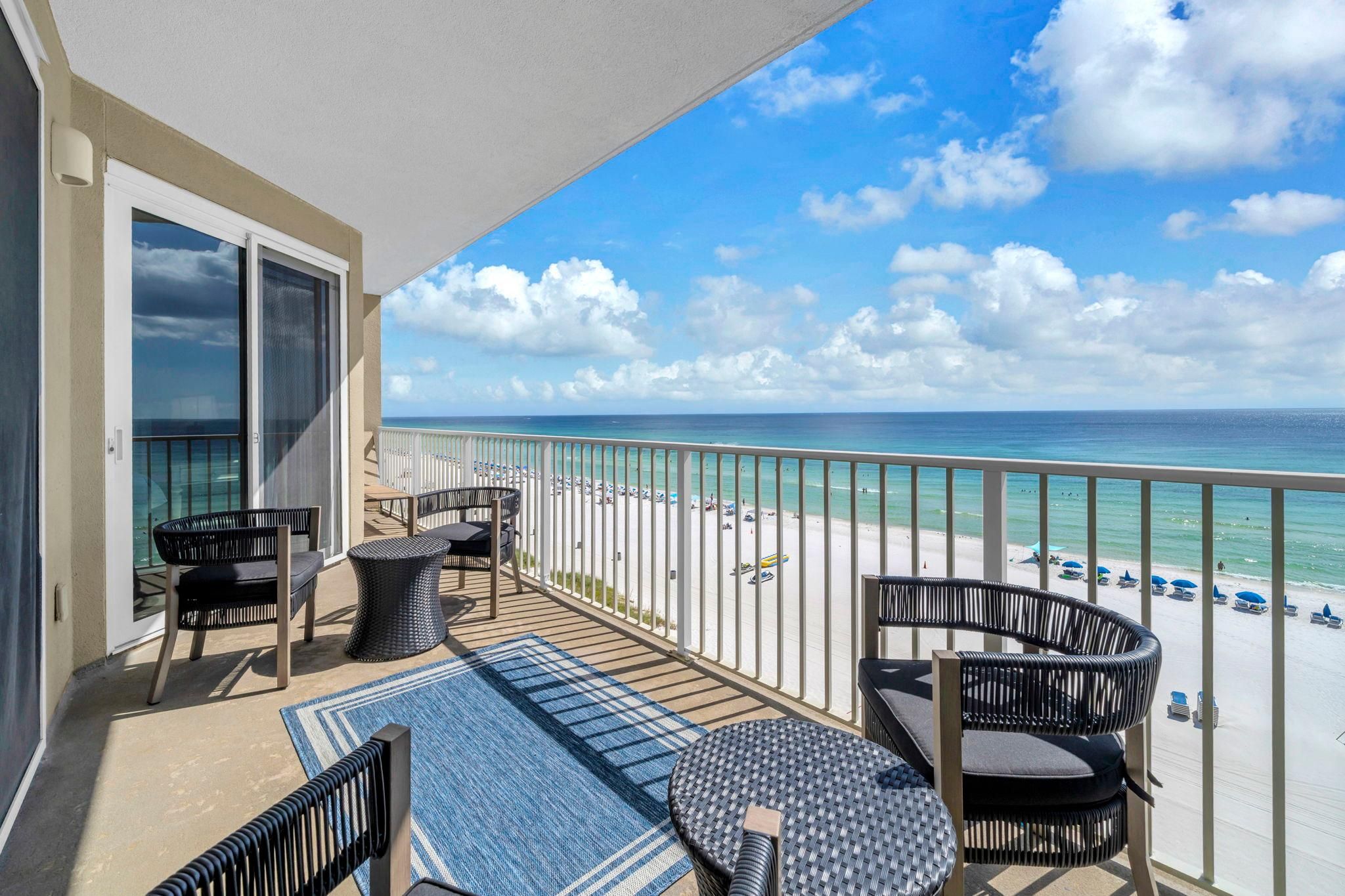 En balkon med udendørs møbler og udsigt til stranden, under en blå himmel.