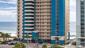 A multi-story hotel with a blue and beige color scheme, located near a beach with visible ocean and palm trees. - Grandview East 302 - Sandscape (Panama City Beach)