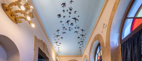 A hallway with a ceiling mural of birds, arched doorways, and a stained glass window.