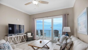 A spacious living room with a flat-screen TV, a wooden coffee table, and a sofa with decorative pillows. There is a balcony with a view of the ocean and a ceiling fan. - Grandview East 1602 (Panama City Beach)