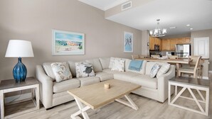 A living room with a beige sofa, a wooden coffee table, and a dining area with wooden chairs and a table. - Grandview East 1602 (Panama City Beach)