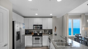 A modern kitchen with stainless steel appliances, white cabinets, granite countertops, and a view of the ocean. - Grandview East 1601 - Azure Paradise (Panama City Beach)
