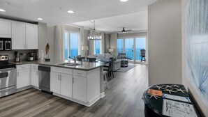 A modern kitchen with white cabinetry, a central island, and a dining area with a view. - Grandview East 1601 - Azure Paradise (Panama City Beach)