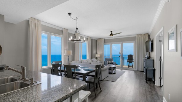 A modern kitchen with granite countertops, a dining area with a table and chairs, a living area with a sofa and a TV, and large windows with a view of the ocean. - Grandview East 1601 - Azure Paradise (Panama City Beach)