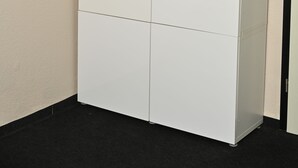 Un armario blanco de cuatro puertas en un rincón de una habitación con paredes color beige y suelo negro.