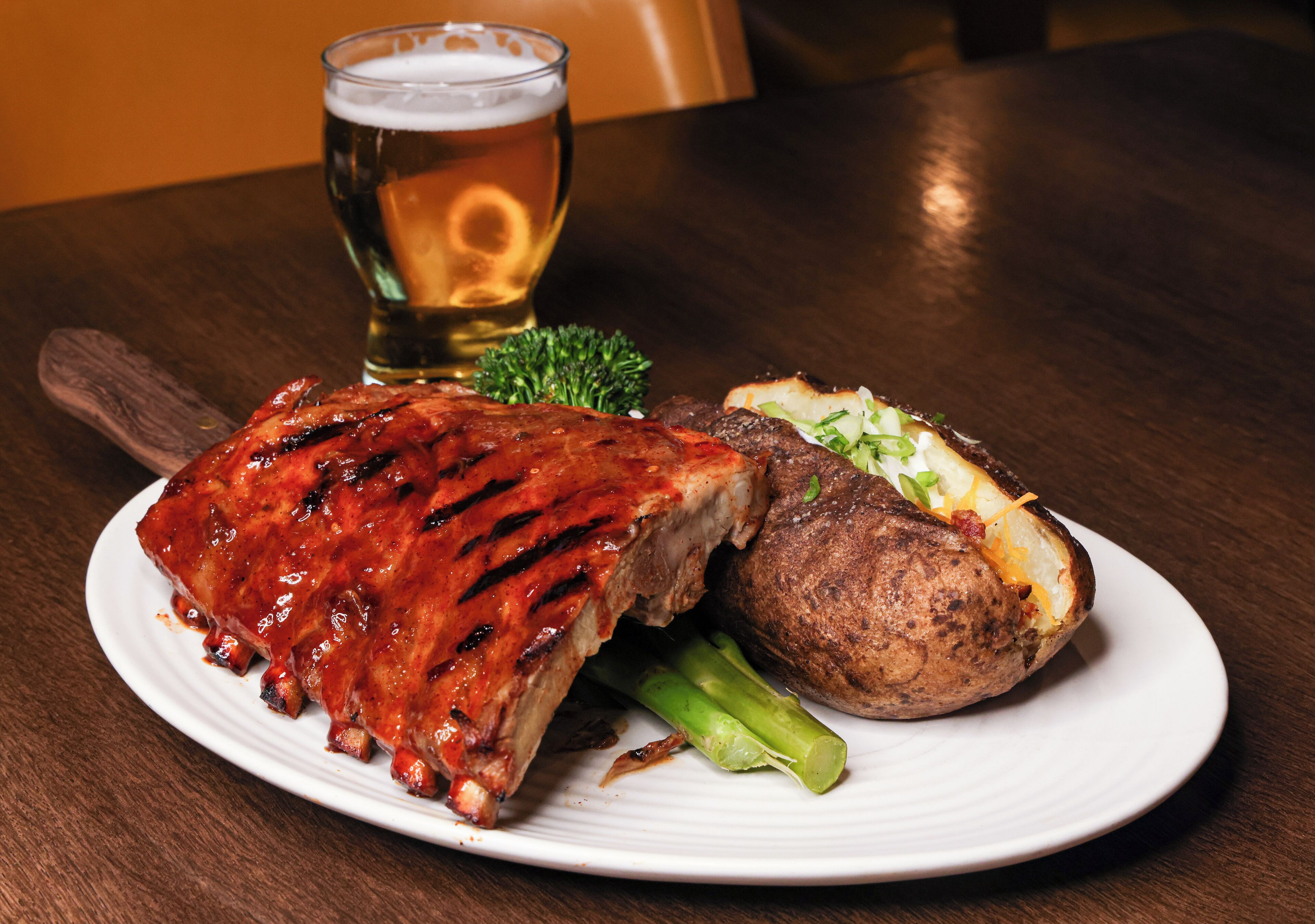 A plate with barbecued ribs, a baked potato, and a glass of beer on a wooden table.