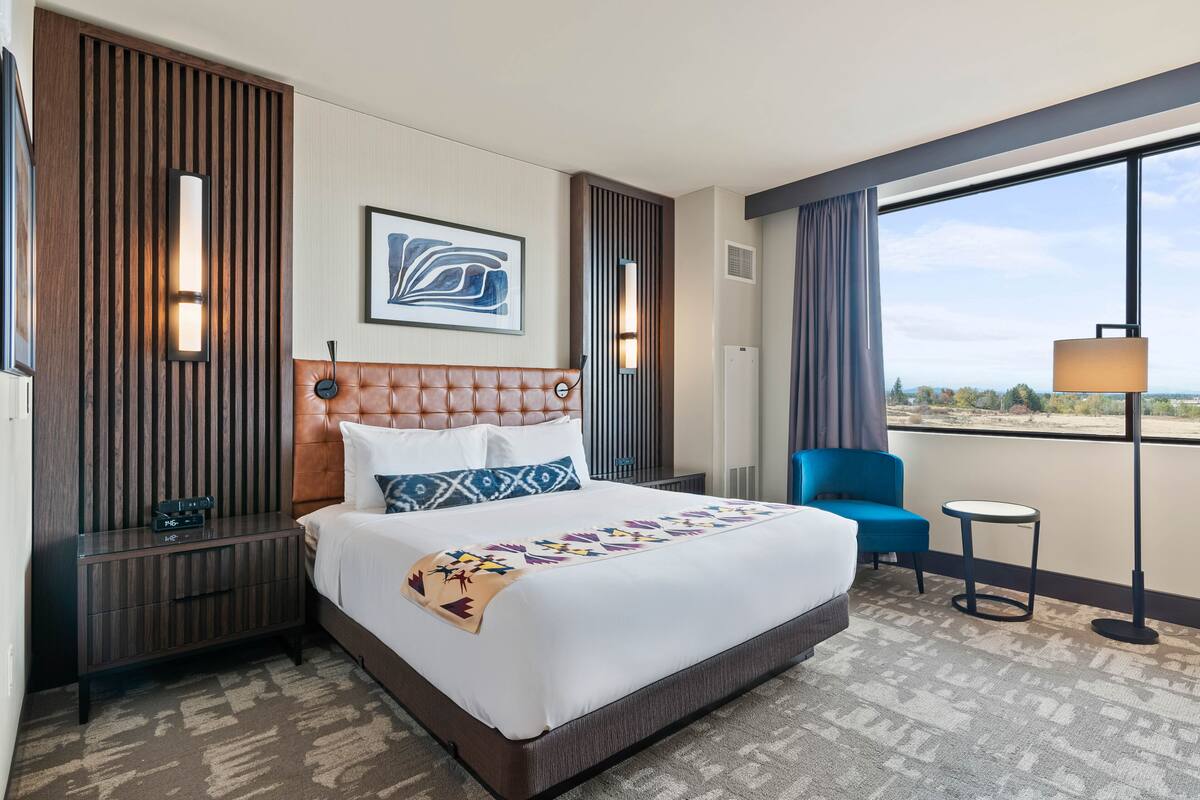 Una habitació d'hotel amb un llit gran, una petitetta, una cadira blava, una taula petita i una finestra gran amb vistes.
