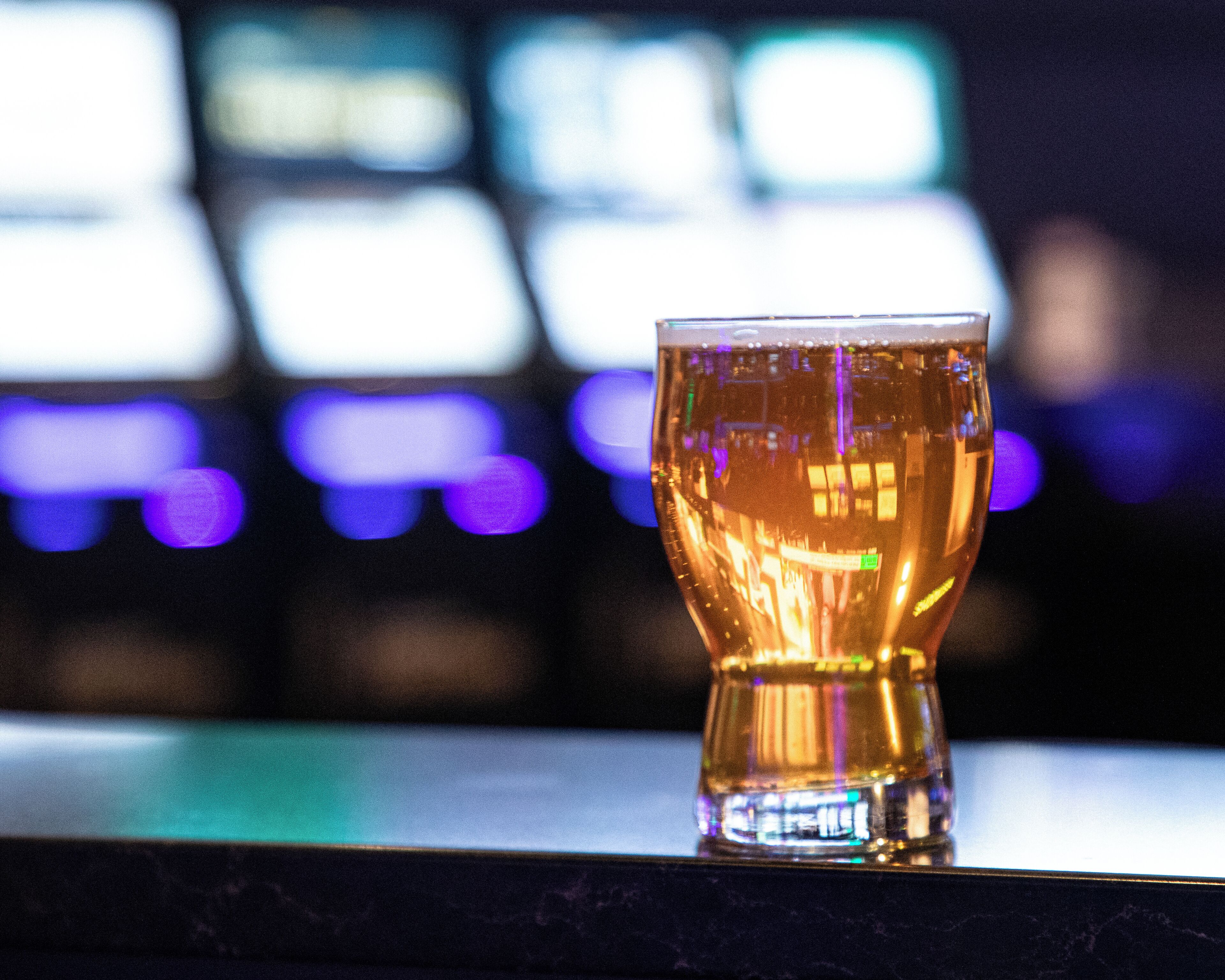 A glass of amber liquid on a reflective surface with a blurred background featuring bright lights.