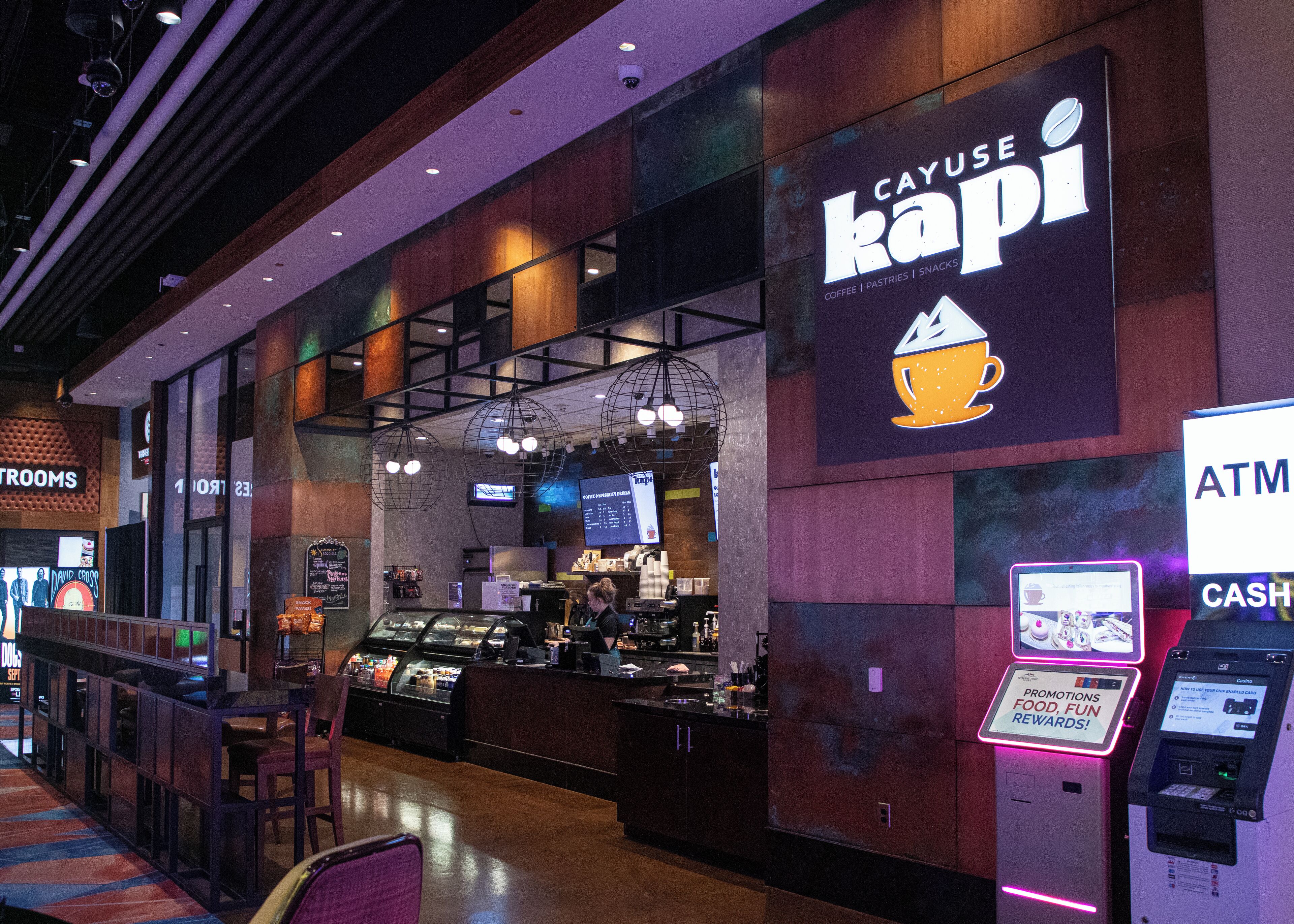 Un café llamado 'CAYUSE KAPI' con un cajero automático y un dispositivo para sacar efectivo a la derecha, una vitrina con pasteles y una zona de estar con sillas.