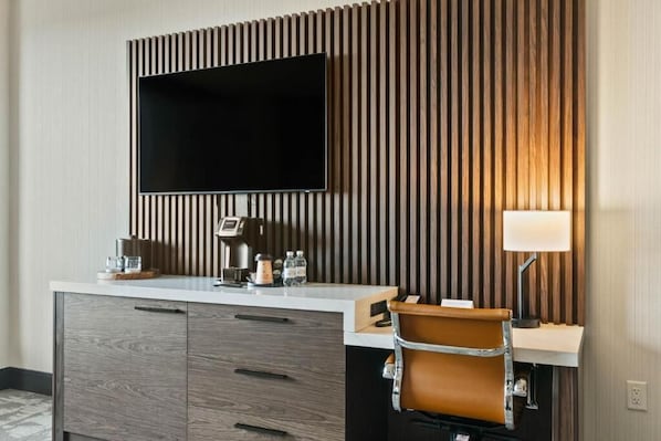 Habitación de hotel moderna con una televisión de pantalla plana instalada en una pared de paneles de madera, un minibár blanco, un sillón de cuero marrón y una mesita de noche con lámpara.