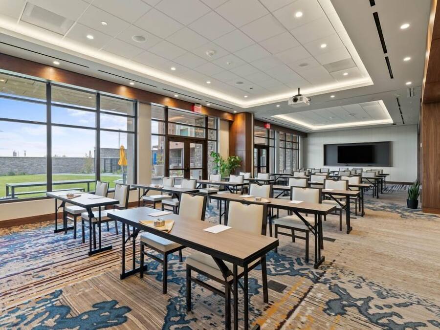 A conference room with multiple tables and chairs, large windows offering a view of the outdoors, and a flat-screen TV mounted on the wall.