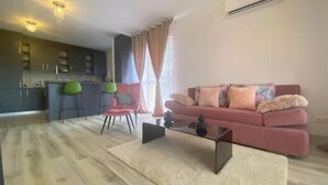 Un salon moderne avec un canapé rose, une table basse en verre et un coin cuisine à l’arrière-plan.