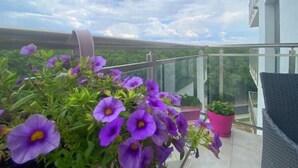 Un balcon orné de fleurs violettes, doté d’une rambarde en verre et offrant une vue sur la verdure.