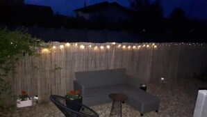 A backyard with a grey sofa, a black chair, and a small table. There are string lights along the fence and potted plants. - Serelor Apartment Free Parking (Dumbrăvița)