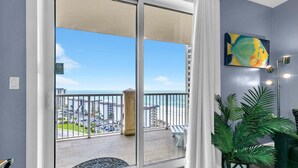 A balcony with a view of the beach and cityscape, a potted plant, and a painting on the wall.