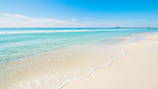 A beach with clear turquoise water, a sandy shore, and a pier in the distance.