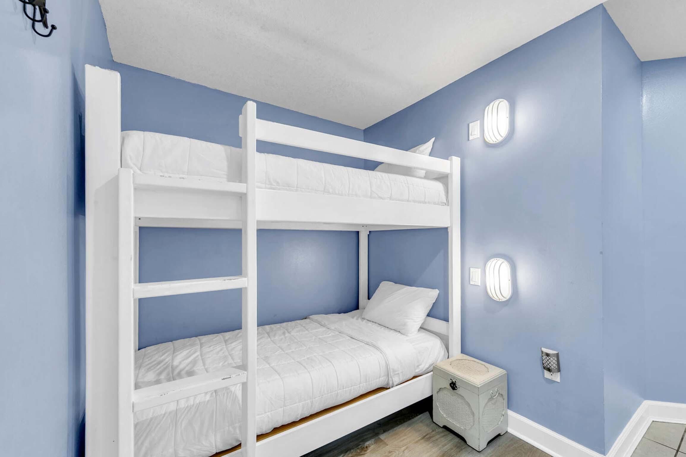 Een stapelbed in een kamer met blauwe muren, wit beddengoed en een klein nachtkastje met een lamp.
