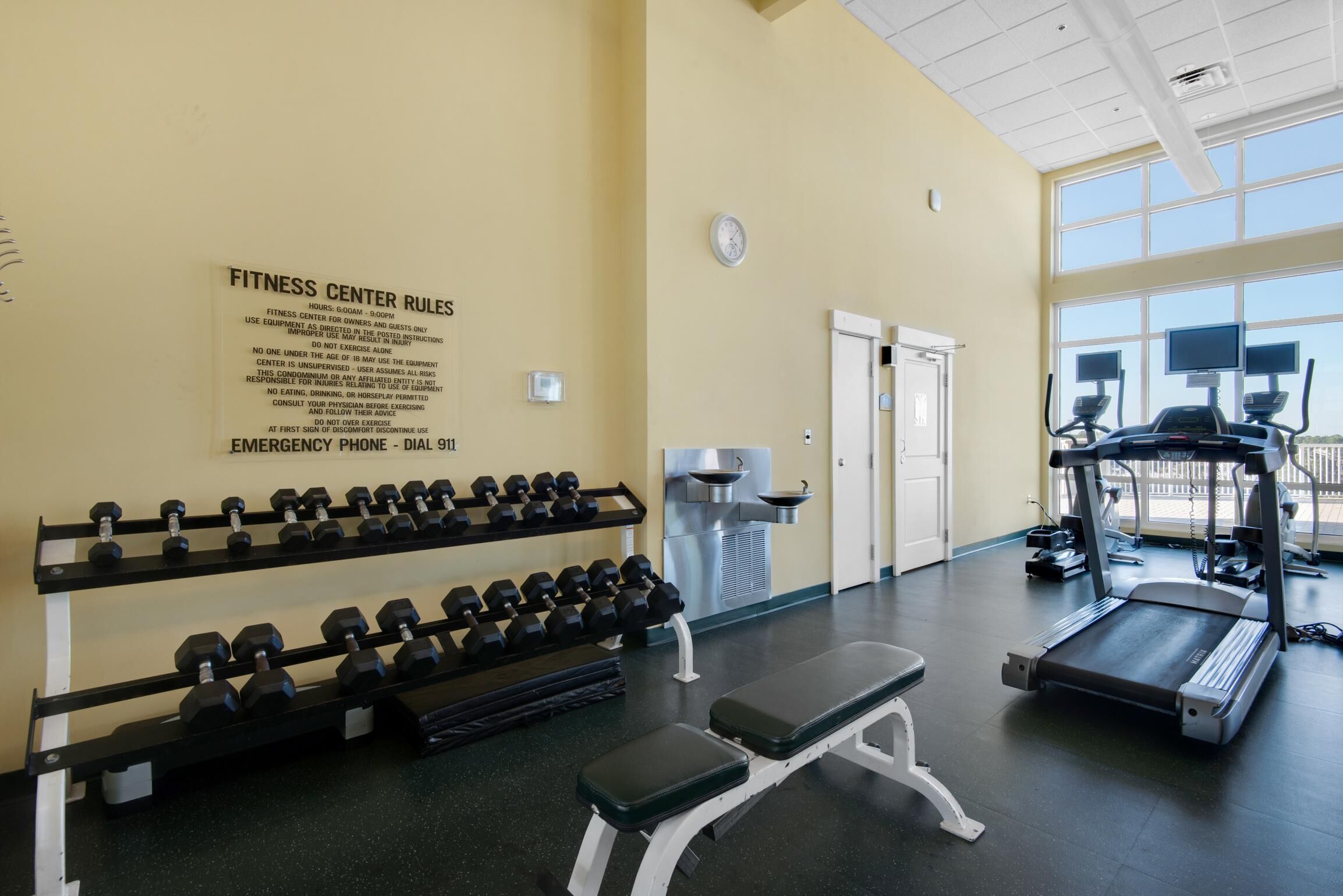 Une salle de sport équipée d’haltères, d’un banc, de tapis de course et d’un panneau mural affichant les règles et les coordonnées à contacter en cas d’urgence.
