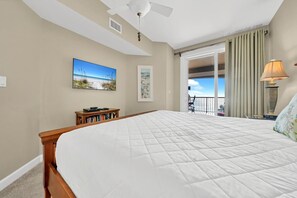 A bedroom with a large bed, a ceiling fan, a view of the beach through an open door, and a nightstand with a lamp. - Grand Panama 1-409 (Panama City Beach)