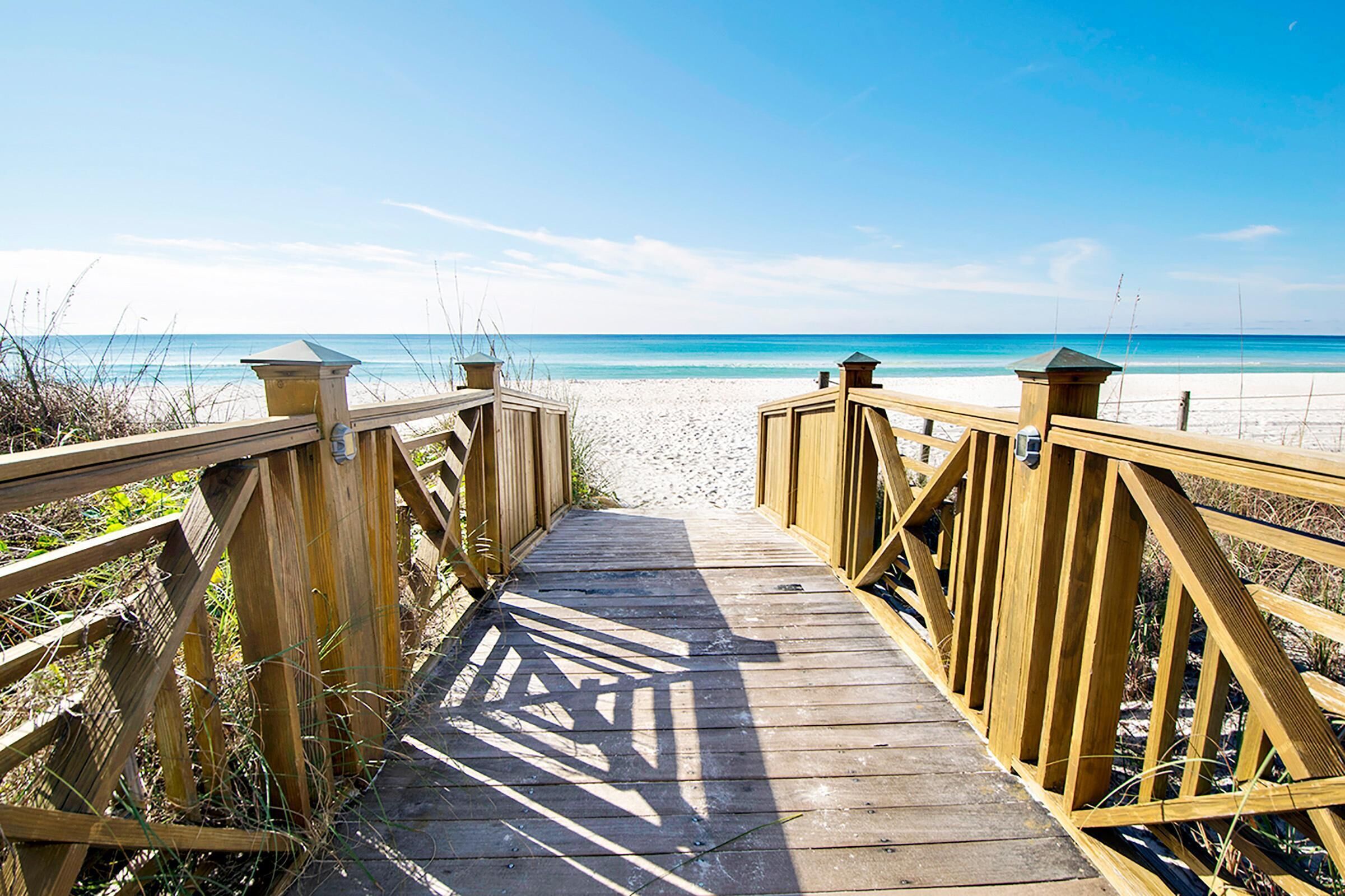 Ein hölzerner Steg führt zu einem Sandstrand mit klarem, blauem Wasser.