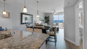 A modern kitchen with granite countertops, a dining area with a white table and chairs, and a view of the ocean through large sliding doors.
