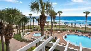 A pool area with lounge chairs and palm trees, overlooking a beach with people and umbrellas.