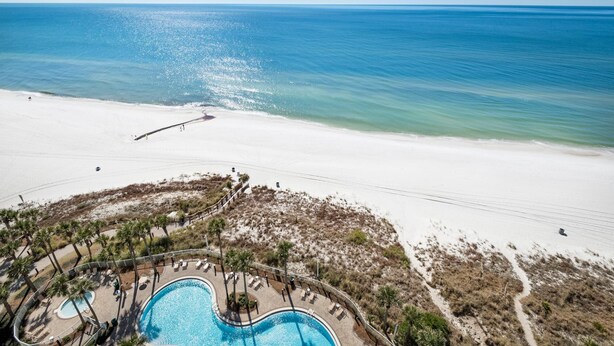 An aerial view of a beach resort with a swimming pool, palm trees, and a clear view of the ocean. - Grand Panama 1-1401 (Panama City Beach)