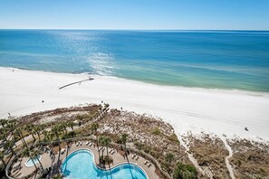 An aerial view of a beach resort with a swimming pool, palm trees, and a clear view of the ocean.