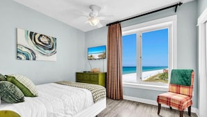 A bedroom with a bed, a green nightstand, a TV, a window with a view of the beach, and a chair with a plaid blanket. - Grand Panama 1-1401 (Panama City Beach)