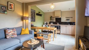 Un salon comprenant un canapé, une table basse et un coin repas avec une table et des chaises en bois. On aperçoit une cuisine à l’arrière-plan, avec un dosseret de crédence en carreaux noirs et des meubles de cuisine en bois.