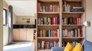 Un salon moderne avec une grande bibliothèque en bois remplie de livres, un canapé gris agrémenté de coussins jaunes et une vue sur la cuisine.