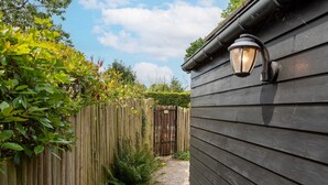 Une clôture en bois, avec un chemin de gravier menant à un bâtiment dont le revêtement extérieur est de couleur sombre et qui est agrémenté d’une lanterne fixée au mur.