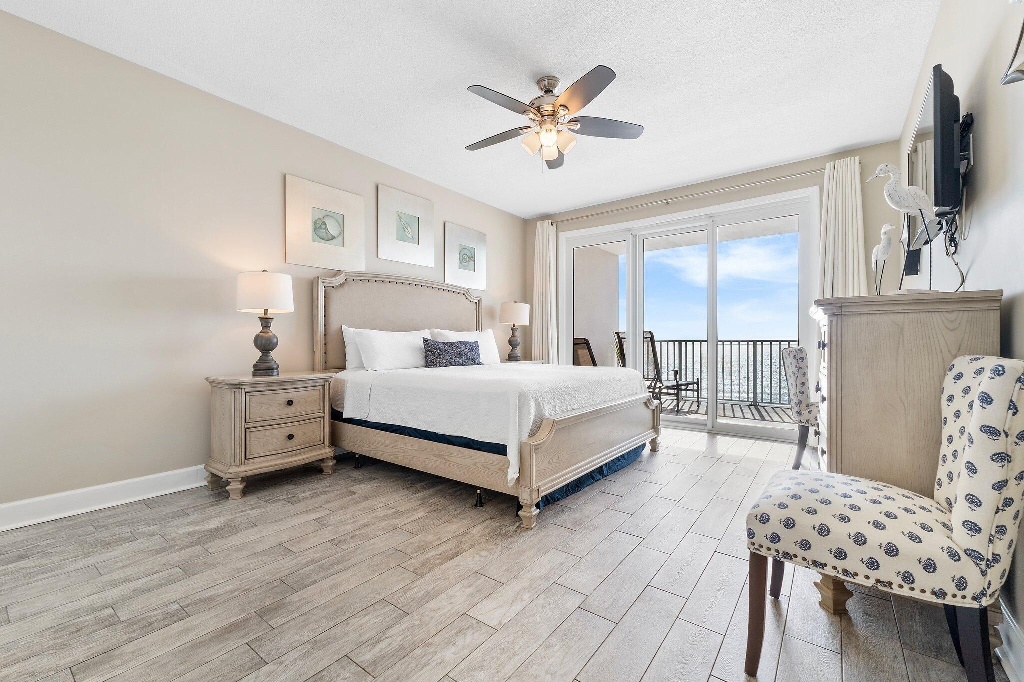 A bedroom with a bed, bedside tables, a chair, a ceiling fan, and a balcony with a view.