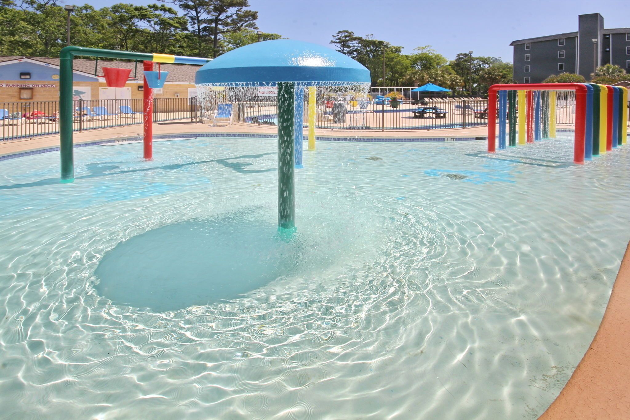 Een waterpretgebied met een fontein in het midden, omringd door een zwembad en een afgesloten zone met ligbedden en parasols.
