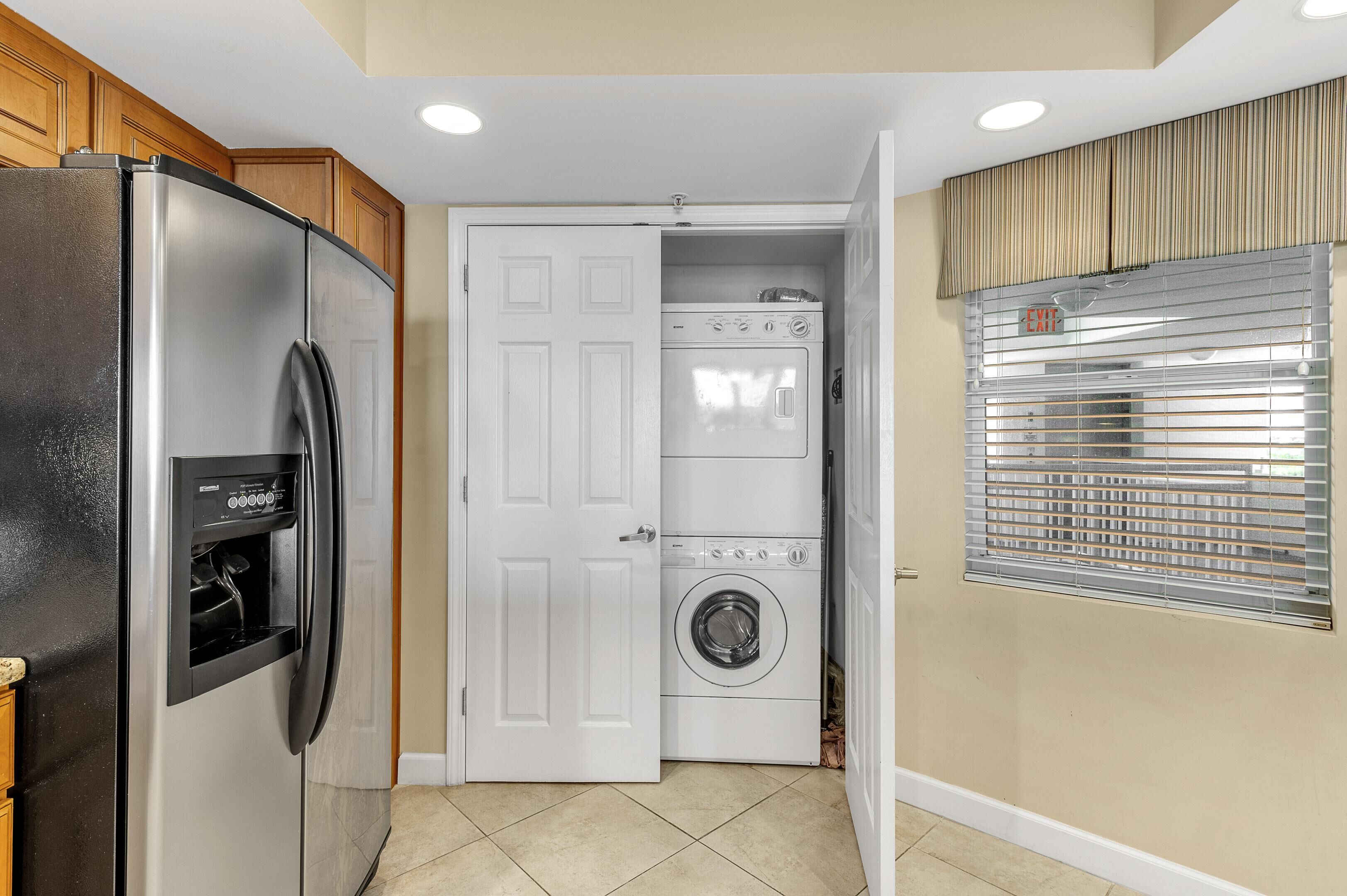 A compact kitchen with a refrigerator, a built-in washer and dryer, and a window with blinds.