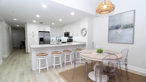 A modern kitchen with white cabinets, a stainless steel refrigerator, and a dining area with a round table and clear chairs. - Spanish Key 603 (Pensacola)