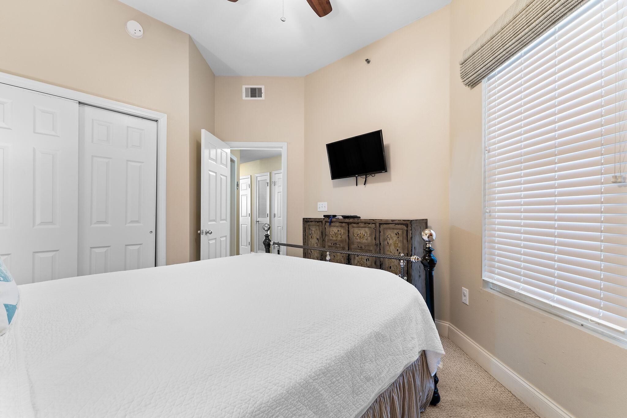A bedroom with a bed, a TV mounted on the wall, a wooden dresser, and a window with blinds.