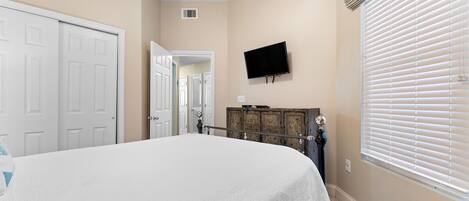 A bedroom with a bed, a TV mounted on the wall, a wooden dresser, and a window with blinds.
