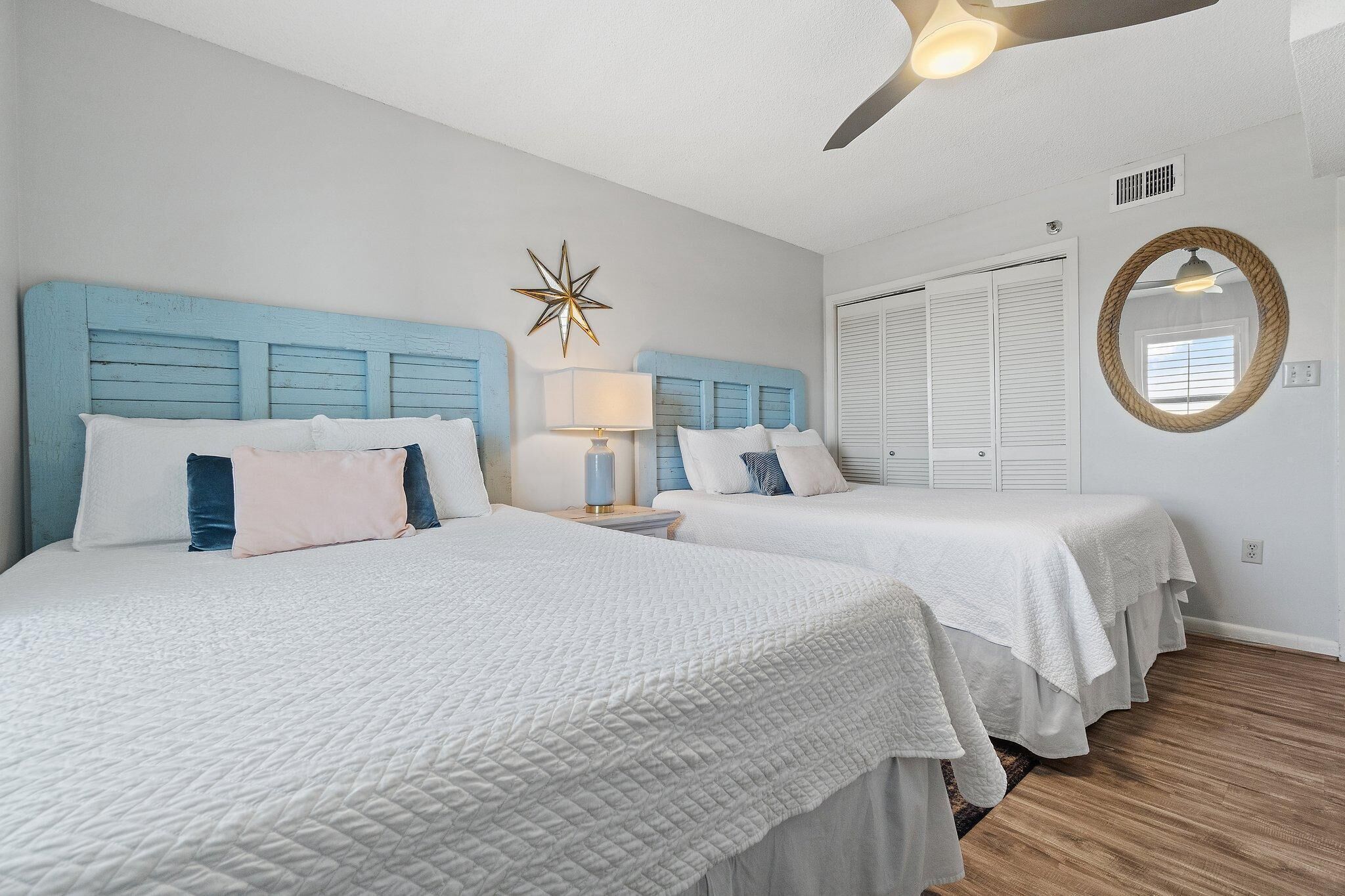 Habitación de hotel con dos camas, ventilador de techo, adorno con forma de estrella, espejo redondo y ventana con persianas.