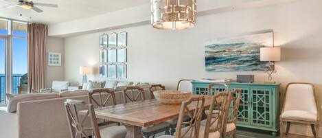 Un coin repas avec une table et des chaises en bois, un ventilateur de plafond, un meuble turquoise et un tableau représentant l’océan accroché au mur.