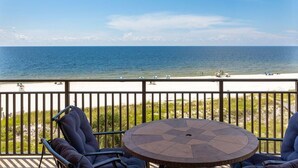 Een balkon met een ronde tafel en twee stoelen, met uitzicht op een strand met mensen en een heldere lucht.