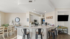A modern kitchen with white cabinets, a central island with bar stools, a clock, and a 'BEACH' sign on the wall. - Seachase 404e (Orange Beach)