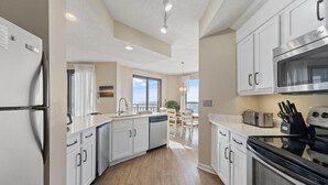 A modern kitchen with white cabinetry, a stainless steel refrigerator, a built-in oven, and a dishwasher. There is a dining area with a view of the ocean. - Seachase 404e (Orange Beach)