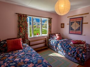 A cozy bedroom with two beds, floral bedding, a large window with curtains, a hanging light fixture, and a quilt on the wall.