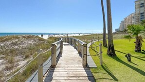 A wooden boardwalk leading to a beach with palm trees and a building in the background. - Summer House 203a (Orange Beach)