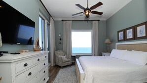 A bedroom with a large bed, a ceiling fan, a TV, a dresser, a chair, and a view of the ocean. - Indigo East 2001 (Pensacola)